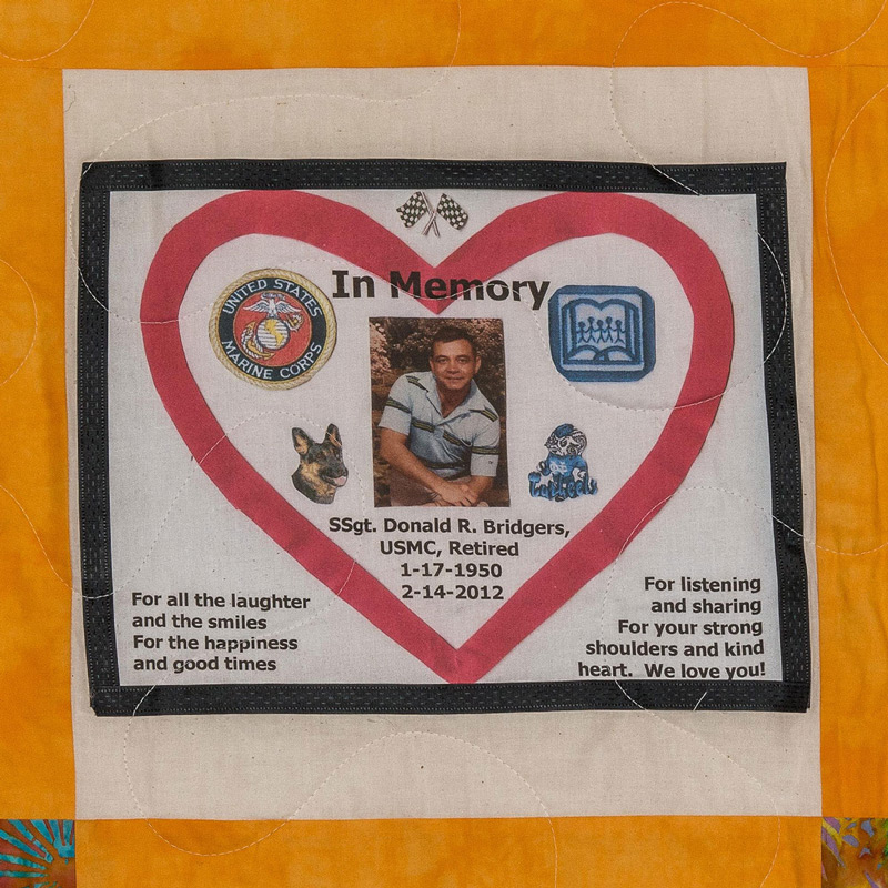 Quilt square for Donald Bridgers with a photo of Donald in the center and patches of racing flags, the marine corps logo, a book, and tarheels mascot. Text reading: for all the laughter and the smiles. For the happiness and good times. For listening and sharing. For your strong shoulders and kind heart. We love you!