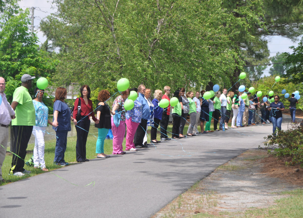 Outdoor Donate Life event with individuals in a line, holding each other’s hands and blue and green balloons