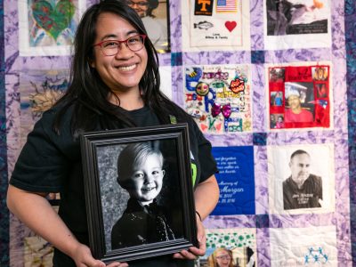 Photo of woman holding her donor's photo in front of a donor quilt