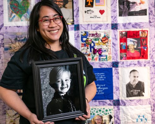 Photo of woman holding her donor's photo in front of a donor quilt