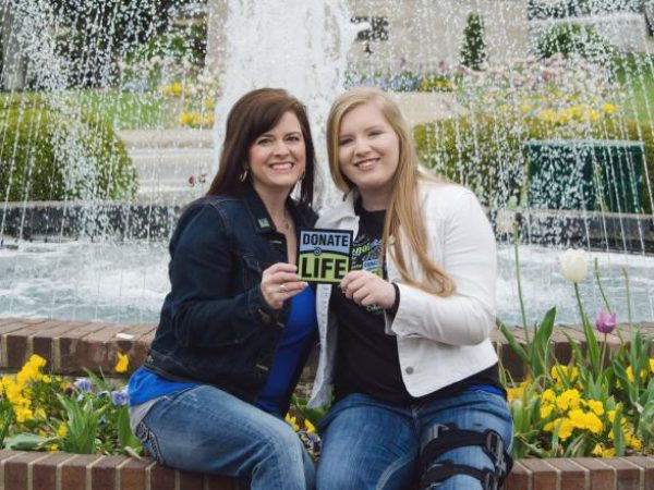 Organ recipient, Mattie, holds Donate Life sign with daughter in front of fountain