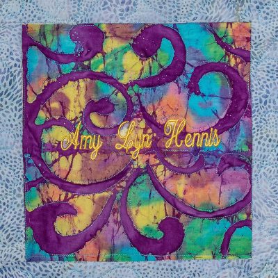 Quilt square for Amy Hennis with a colorful purple, blue, green, and yellow pattern.