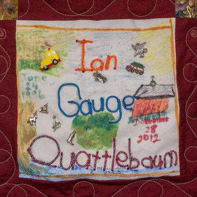 Quilt square for Ian Gauge Quattlebaum with small metal symbols of a cowboy hat, horse, saddle, boots, and bird. And features a colorful scene of a house and land.