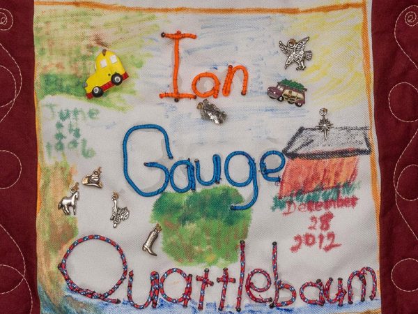 Quilt square for Ian Gauge Quattlebaum with small metal symbols of a cowboy hat, horse, saddle, boots, and bird. And features a colorful scene of a house and land.