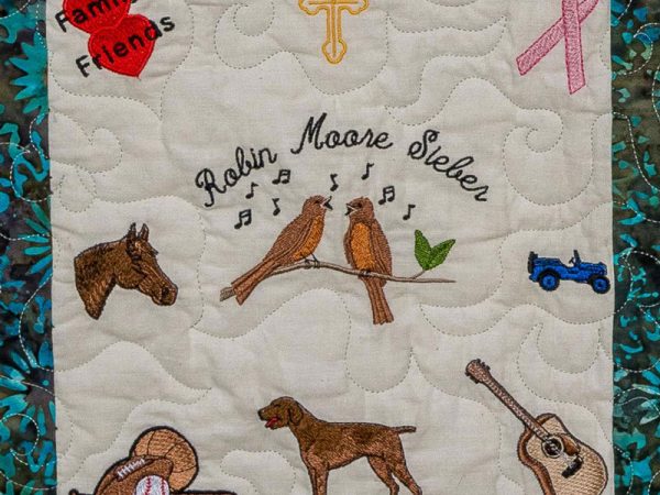 Quilt square for Robin Sieber with patches of family and friends, a cross, birds, a horse, a dog, sports, a guitar, a jeep, and a pink awareness ribbon.
