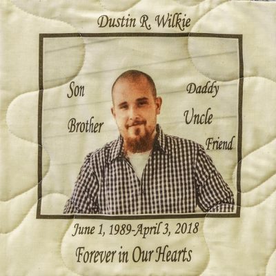 Quilt square for Dustin Wilke with a portrait of Dustin and text reading: Son, Brother, Daddy, Uncle, Friend, Forever in Our Hearts